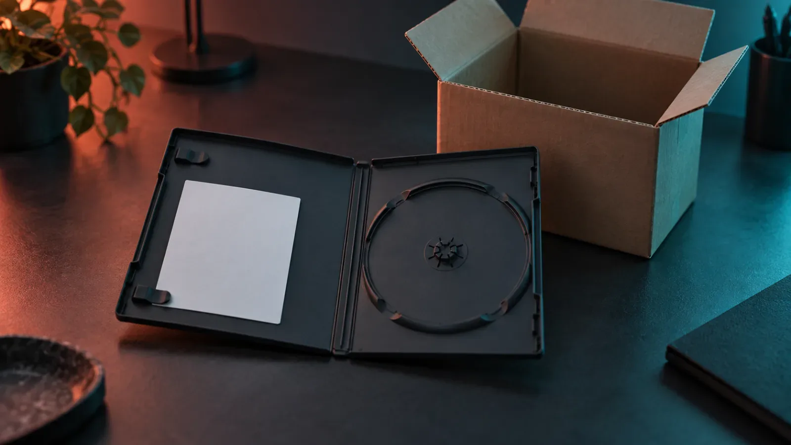 An open, empty black game case on a dark desk, a blank white card in one half and an empty disc holder in the other, with an open empty cardboard box behind